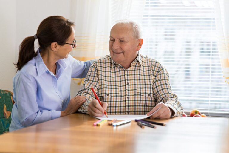 A caregiver sits with an older adult, gently guiding them as they draw with markers at a wooden table near a bright window.