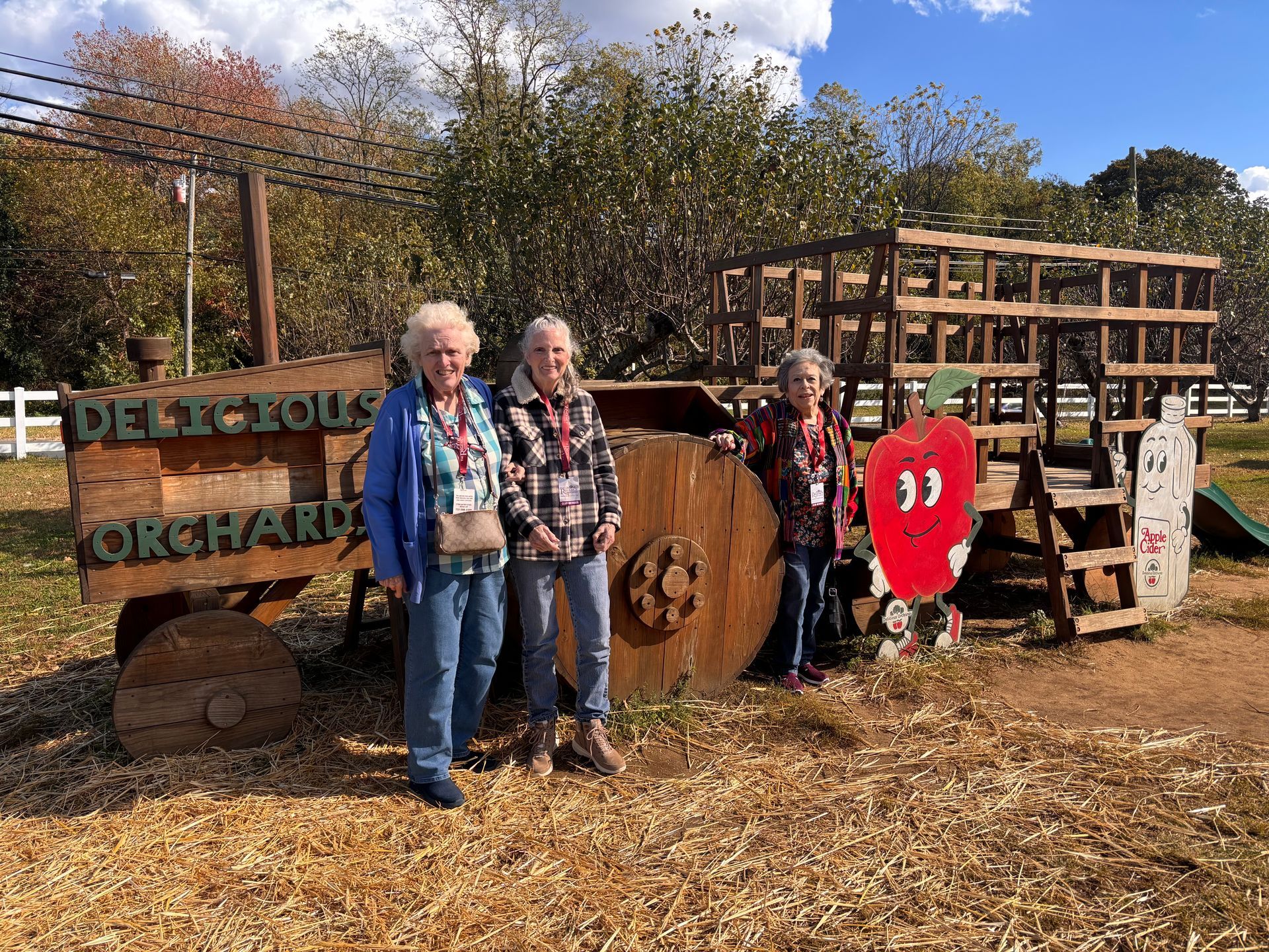 Three people pose in front of a wooden tractor display at an orchard, with a cartoon apple cutout nearby.