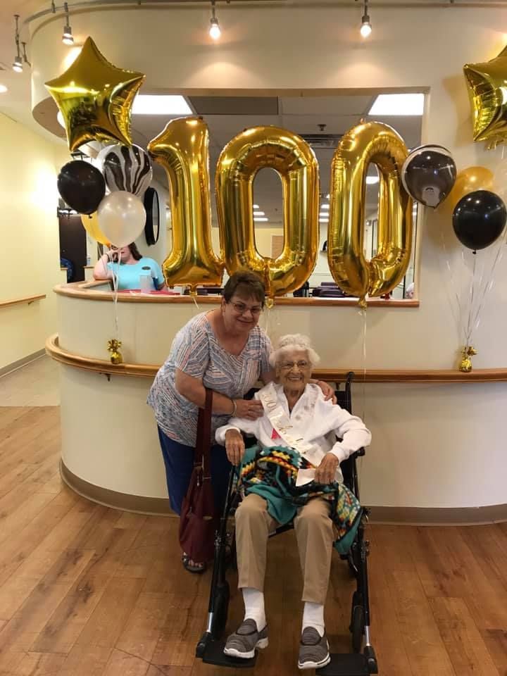A smiling woman stands behind a person in a wheelchair, both posing in front of large gold "100" balloons at a party.