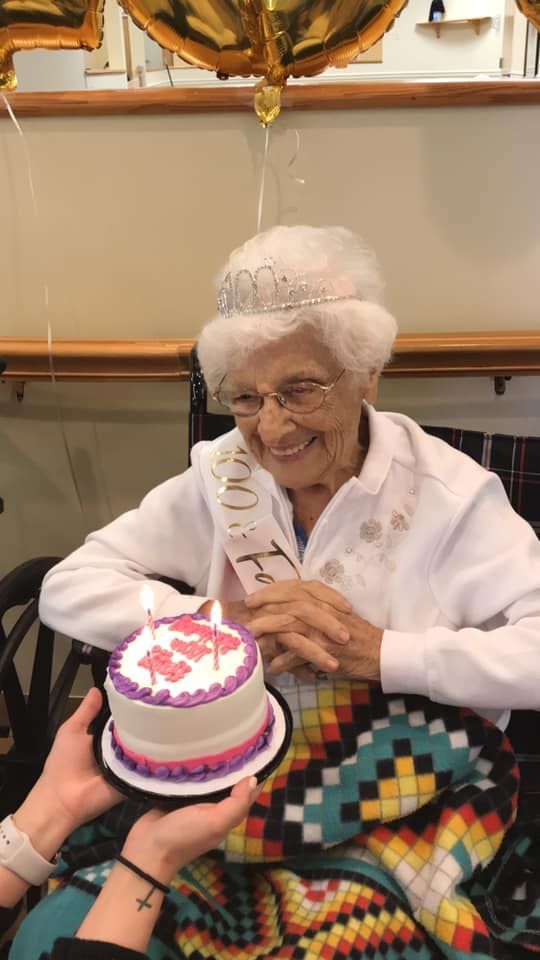 A person smiles while being presented with a small birthday cake with two lit candles.