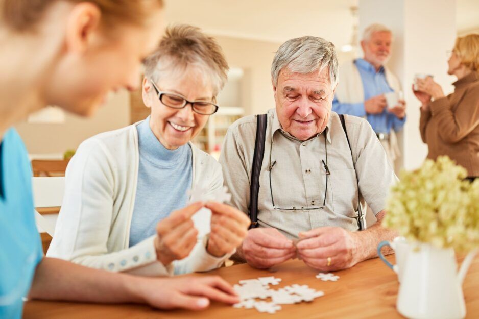 A caregiver and two residents sit at a table working on a puzzle, while others socialize in the background of a room.
