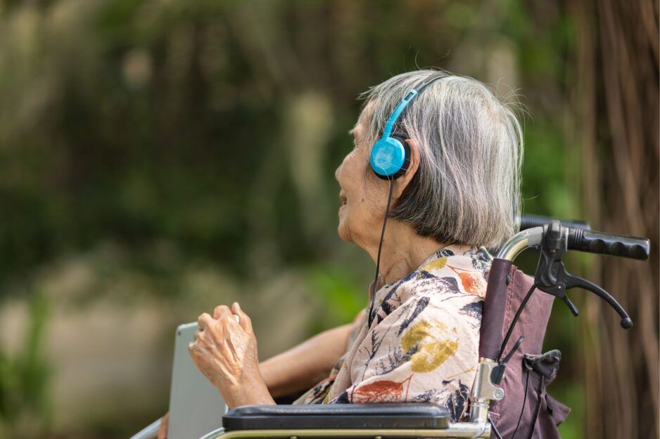 An individual with gray hair sits in a wheelchair outdoors, wearing blue headphones and holding a tablet.