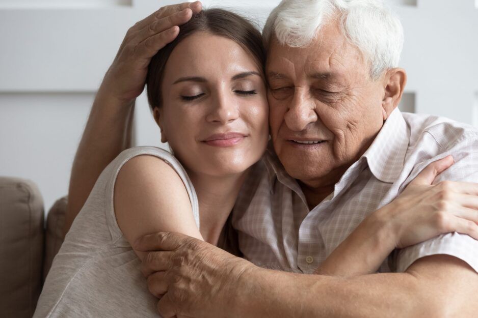 An adult and an elderly person embracing warmly with their eyes closed, resting their heads together.