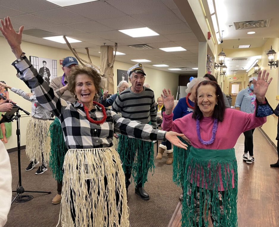 A group of people wearing grass skirts and leis pose in a hallway, smiling with their arms raised as if performing.