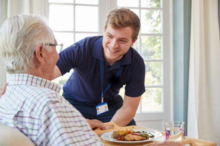 A caregiver in a navy polo shirt smiles while serving a meal on a wooden tray to a person sitting in a chair.