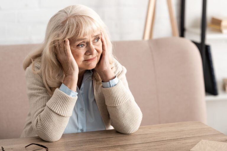 A person with blonde hair rests their head in their hands, looking away with a concerned expression while seated at a table.