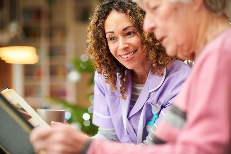 A caregiver in a lilac uniform smiling while looking at a book with an individual in a pink shirt.