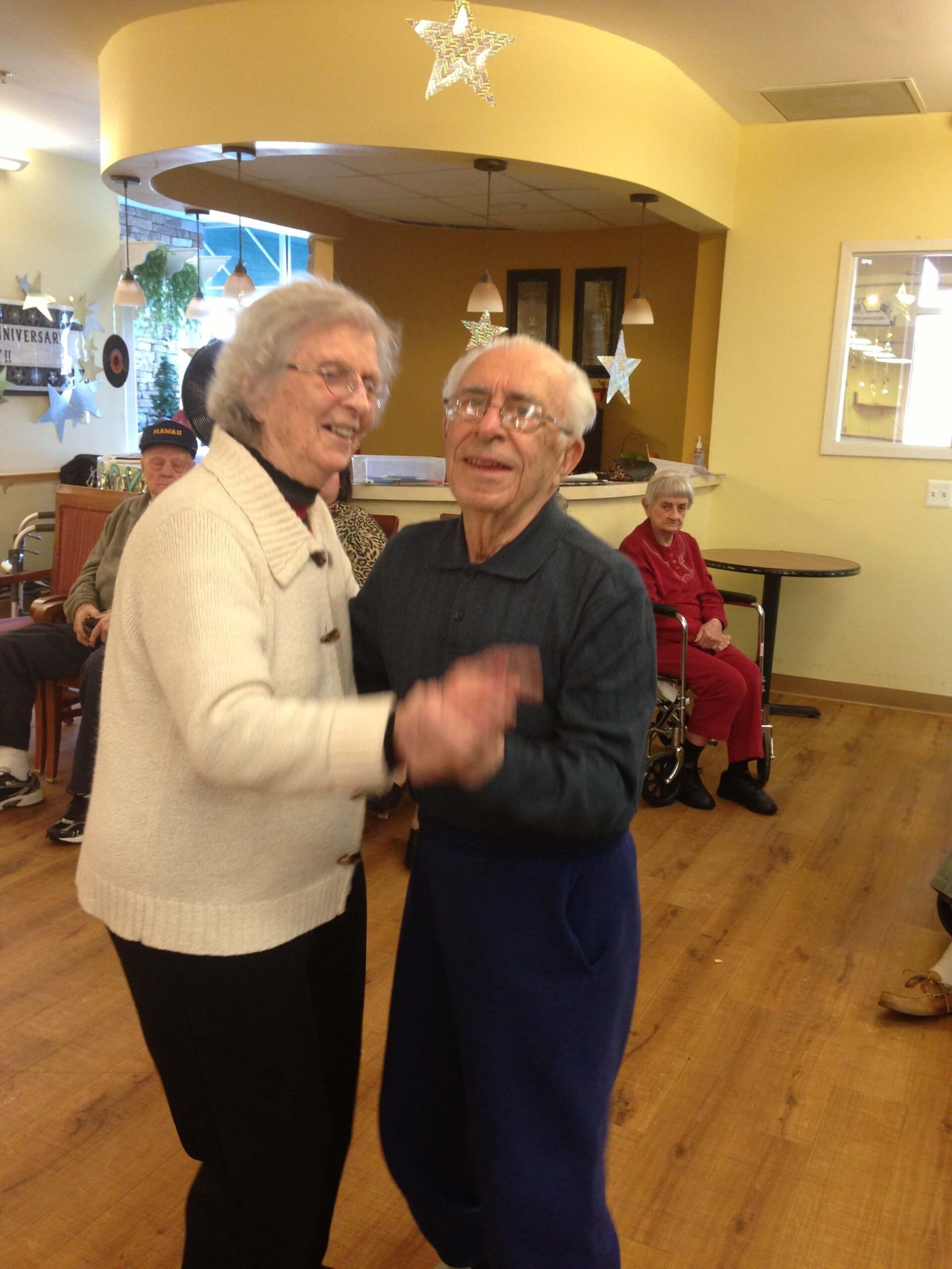 Two people in casual clothing smiling while dancing in a room with light-colored walls and wood-patterned flooring.