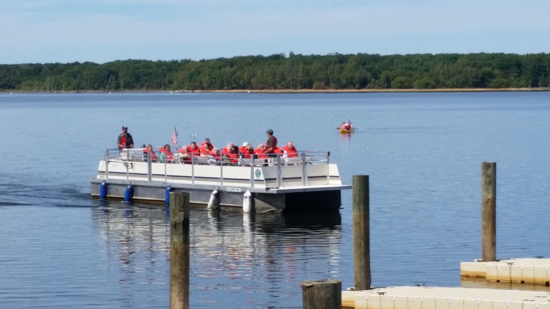 A white pontoon boat carrying a group of people wearing life vests travels across a calm lake near a wooden dock.