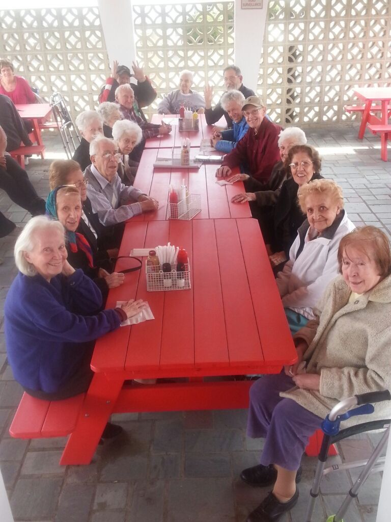 A group of people sitting at a long red picnic table on a patio, some looking at the camera.