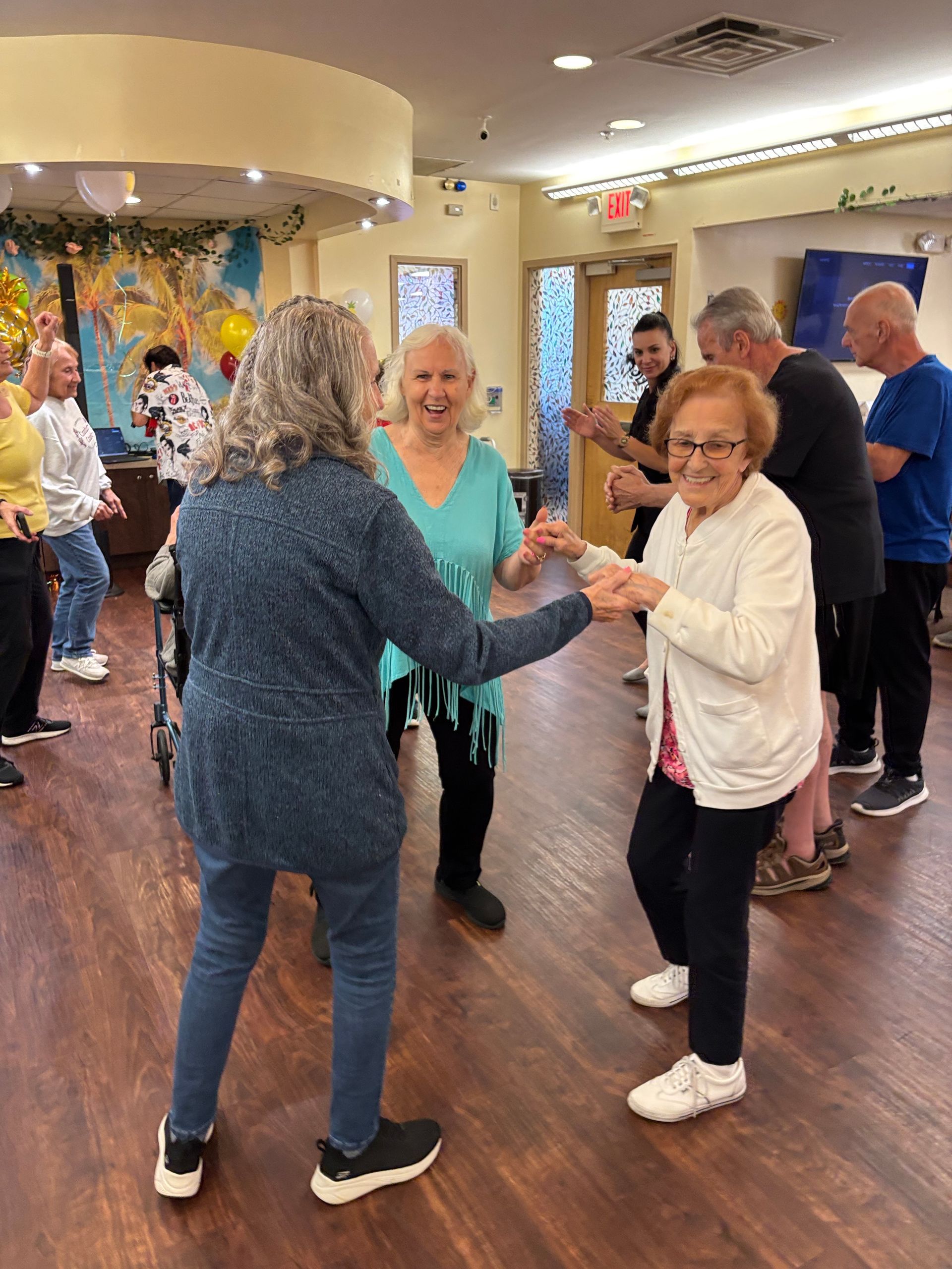 A group of seniors dancing and smiling together in a brightly lit community room with wood-look flooring.