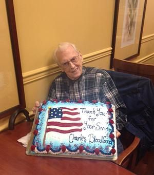 An older person sitting at a table smiling while holding a sheet cake decorated with an American flag and text.