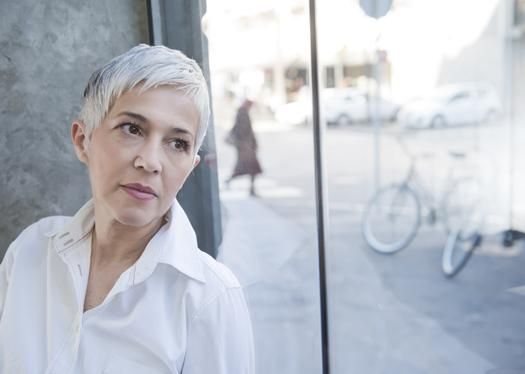 A person with short light hair wearing a white shirt gazes out a glass window at a street scene with a bicycle.