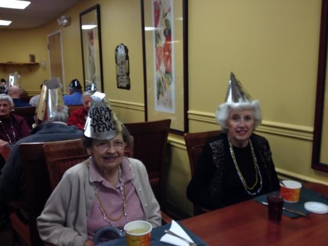 Two smiling people wearing silver New Year's party hats sit at a table in a dining room.