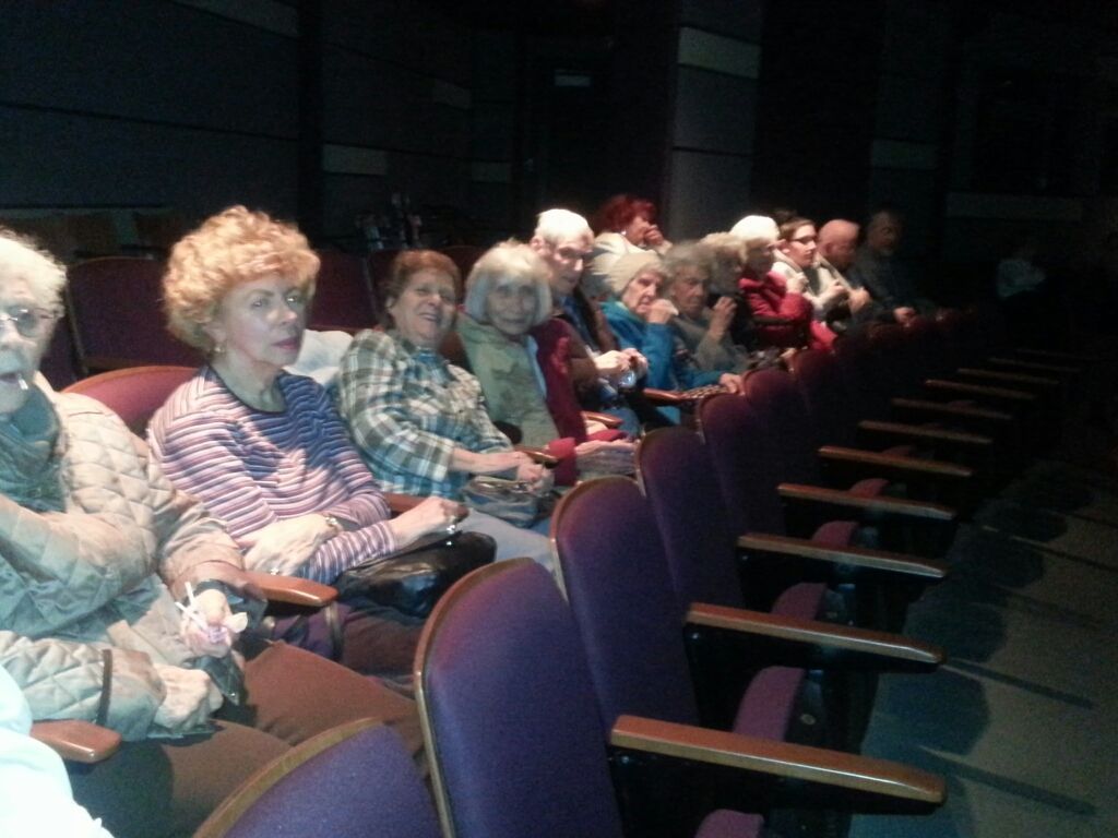 A group of people sitting in rows of theater seats in a dim, indoor venue, looking toward the front.