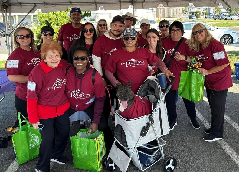 A group of people wearing matching maroon charity shirts, some holding green bags, with a dog in a stroller outdoors.