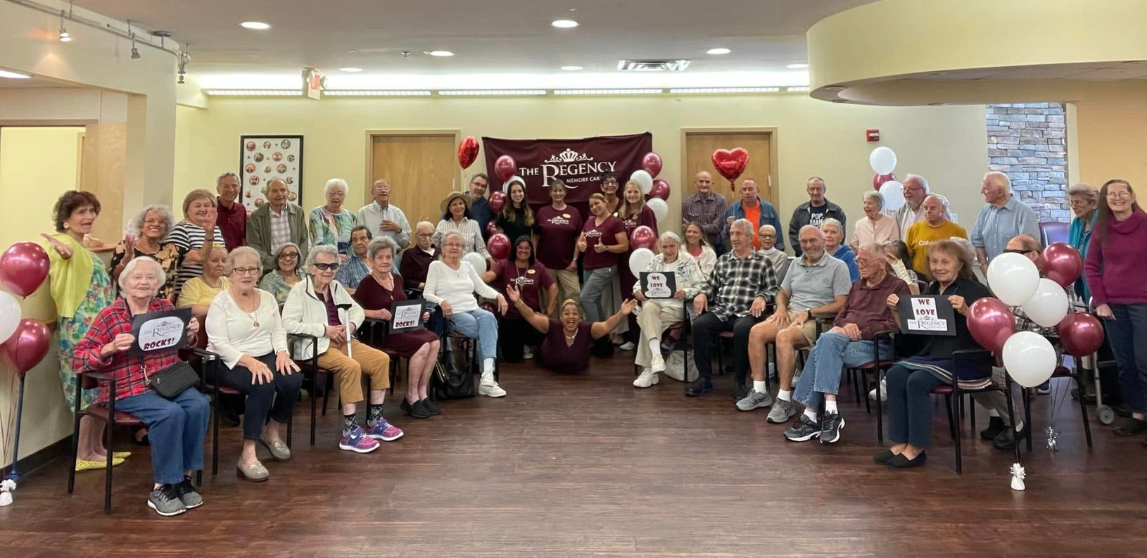 A large group gathers in a community room for a party with maroon and white balloons and decorations.