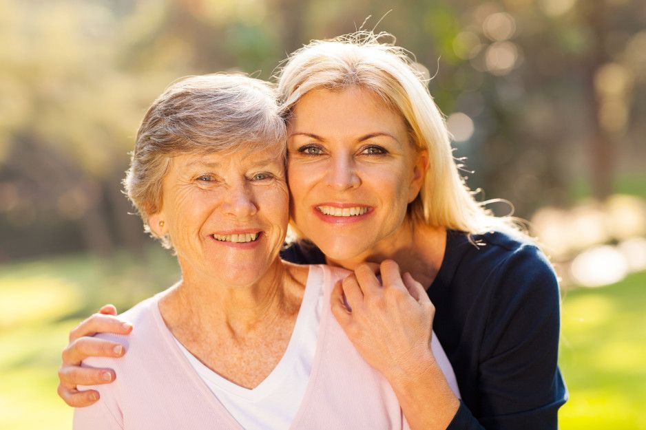 Two smiling people outdoors, one with an arm around the other's shoulder against a soft, sunlit background.