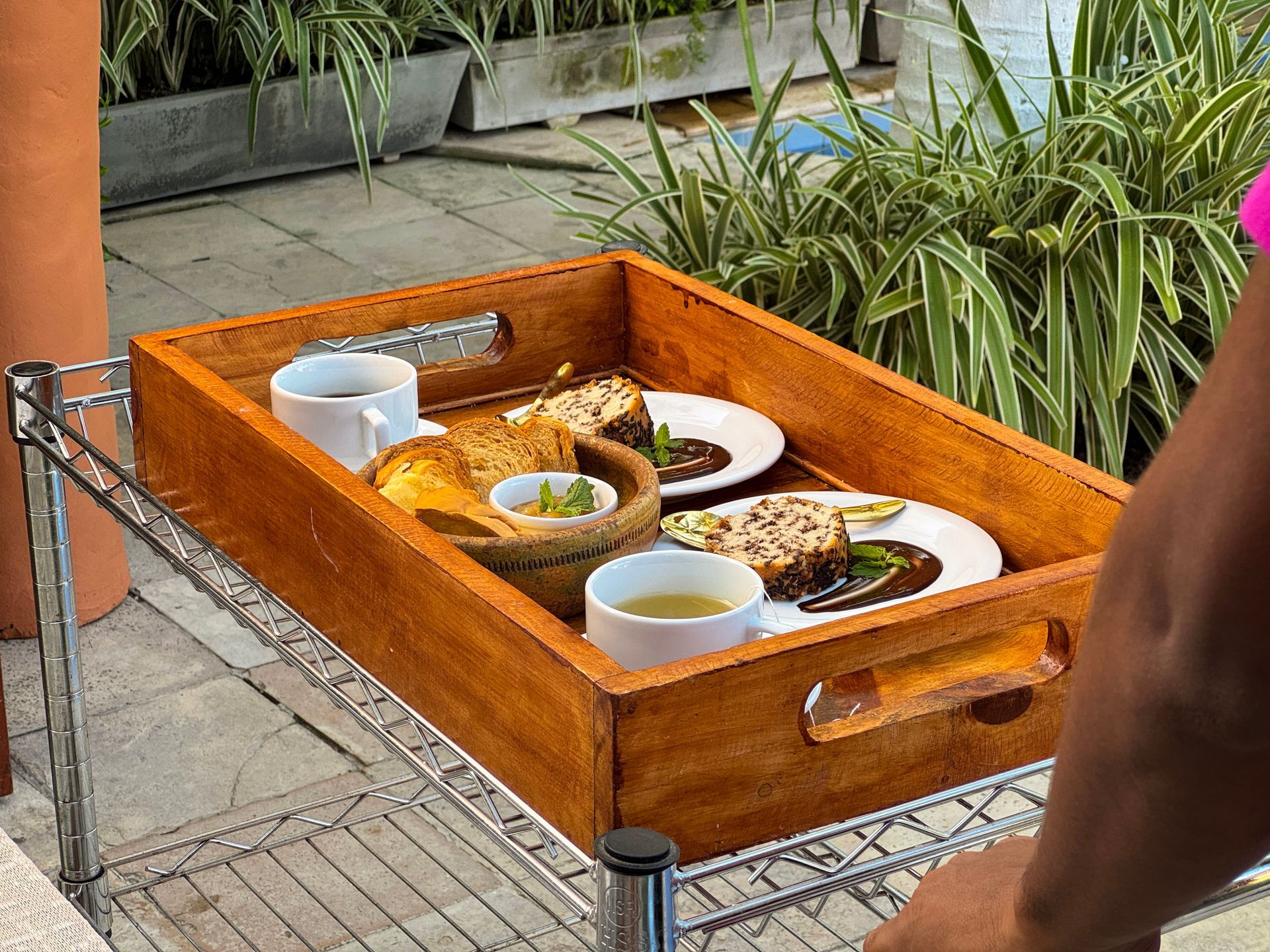 Bandeja com pratos e copos em um carrinho com rodinhas ao ar livre, ao lado de plantas.
