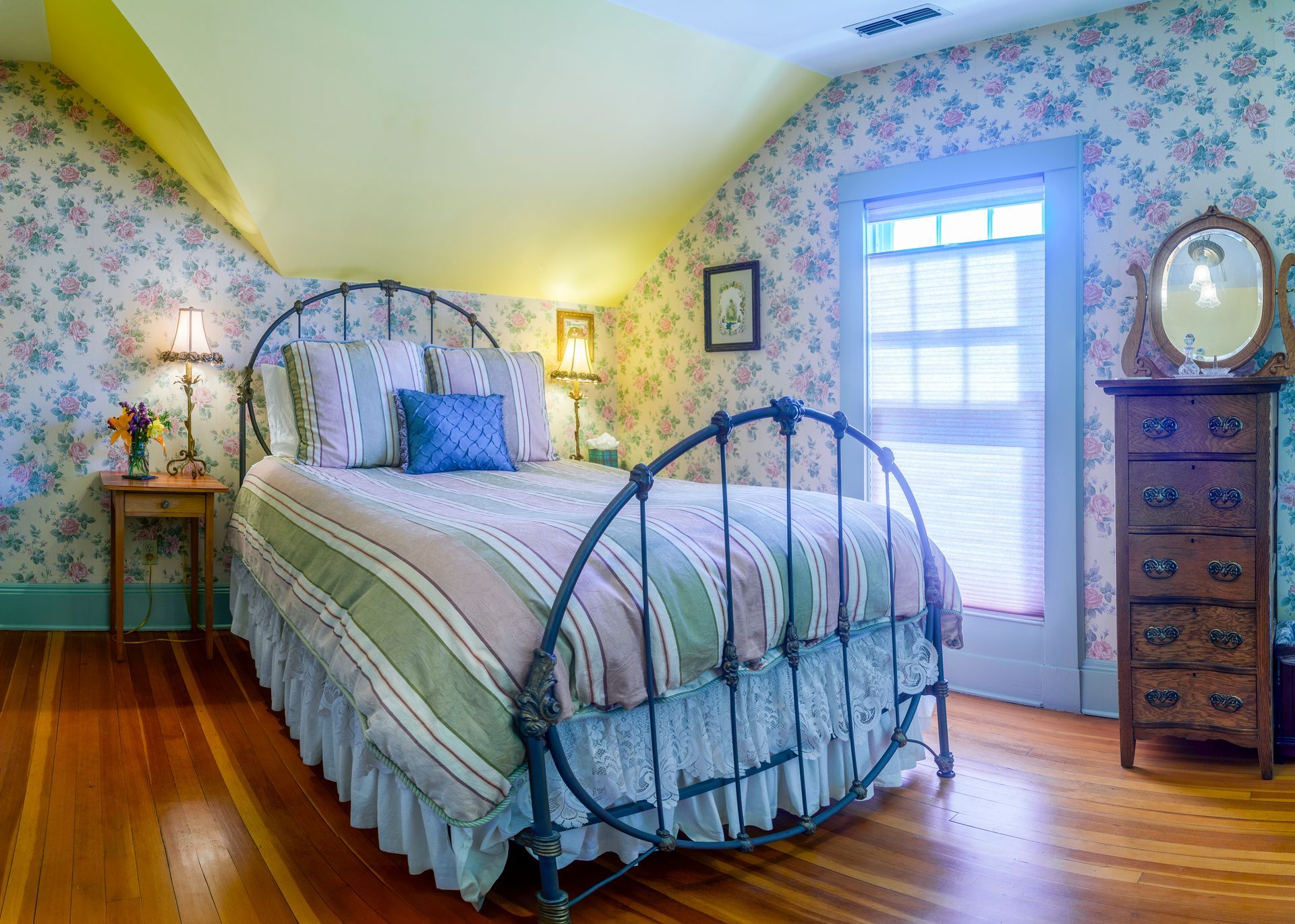 A bedroom with a bed , dresser , mirror and floral wallpaper.