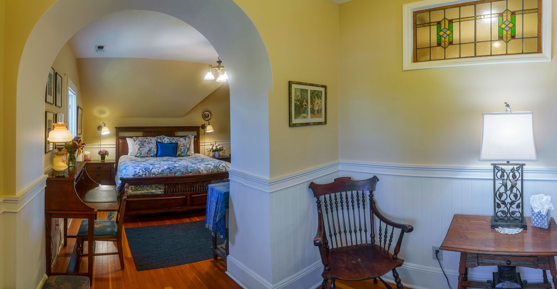 A bedroom with a bed , chair , table , lamp and stained glass window.