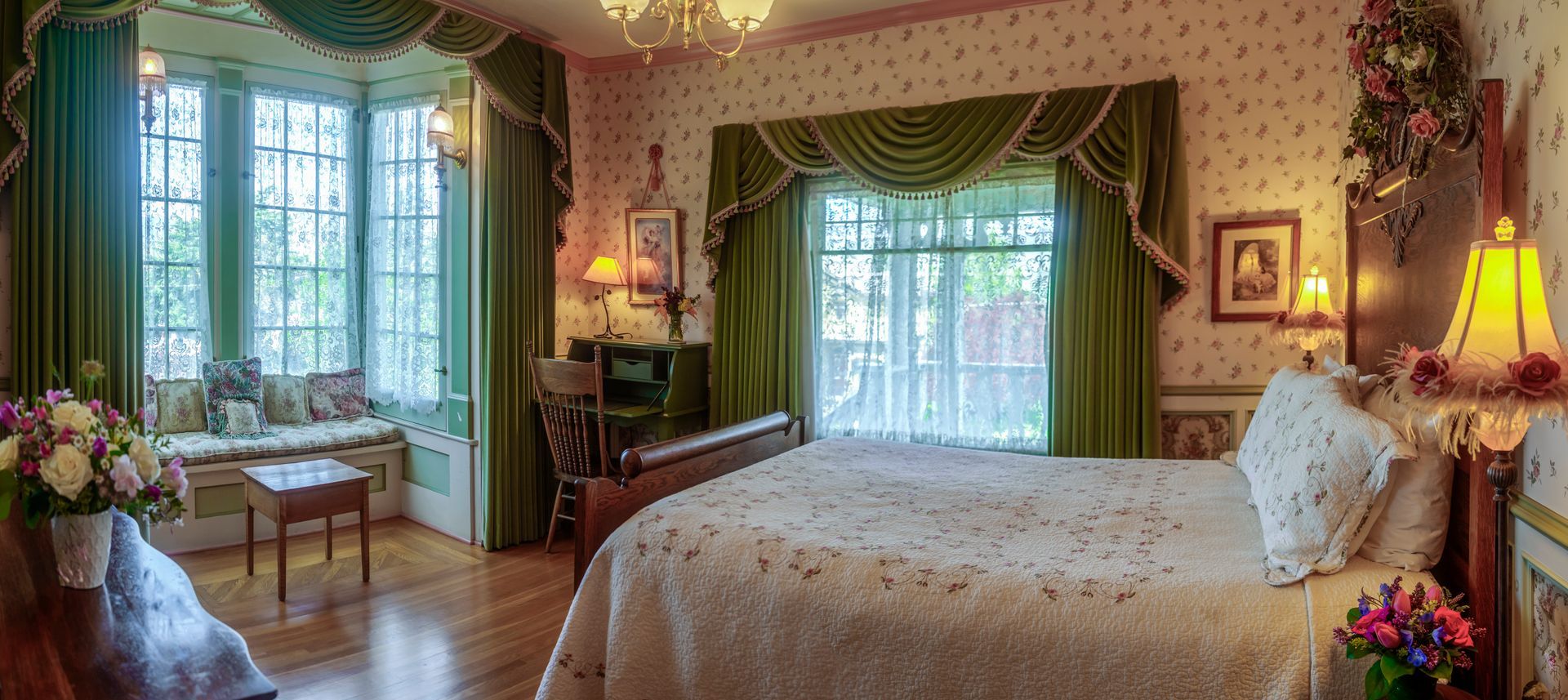 A bedroom with a large bed , green curtains , a desk , lamps , and a large window.