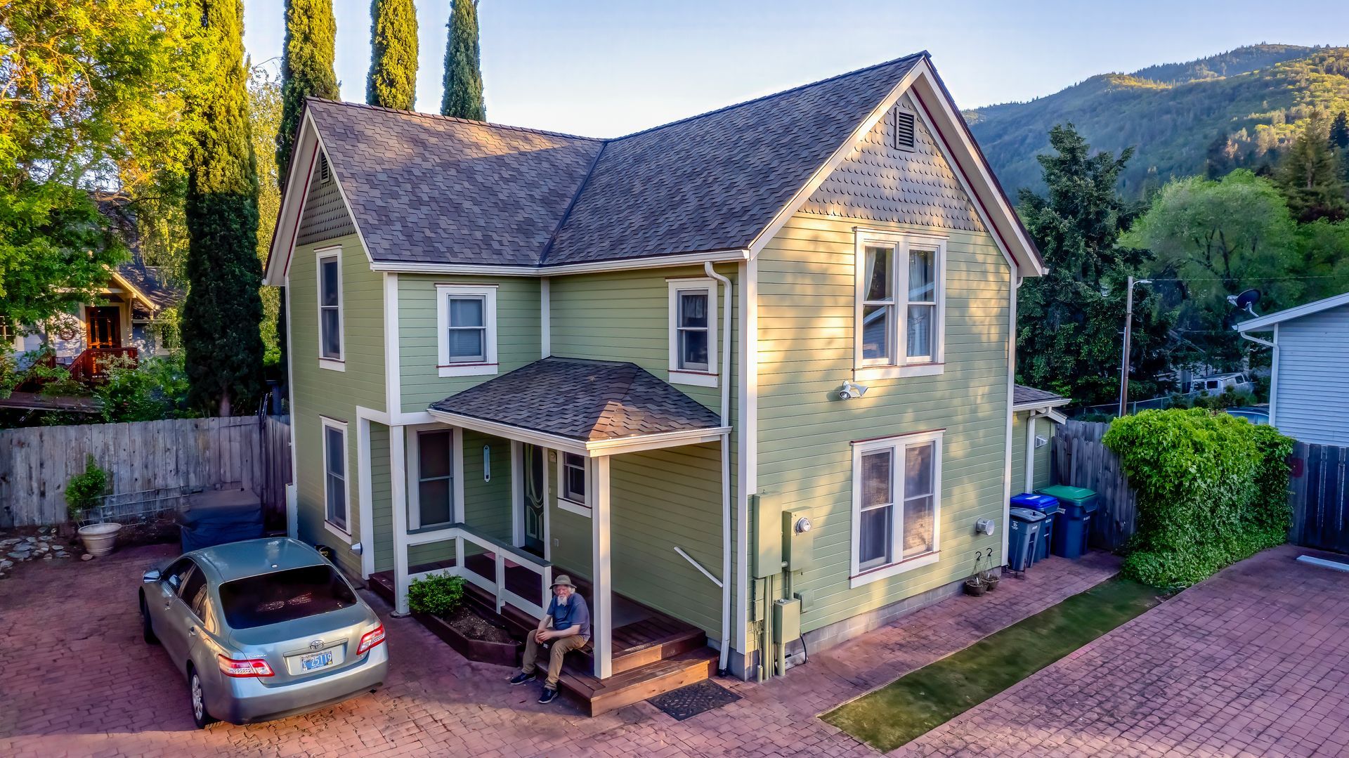 A man is sitting on the porch of a green house with a car parked in front of it.