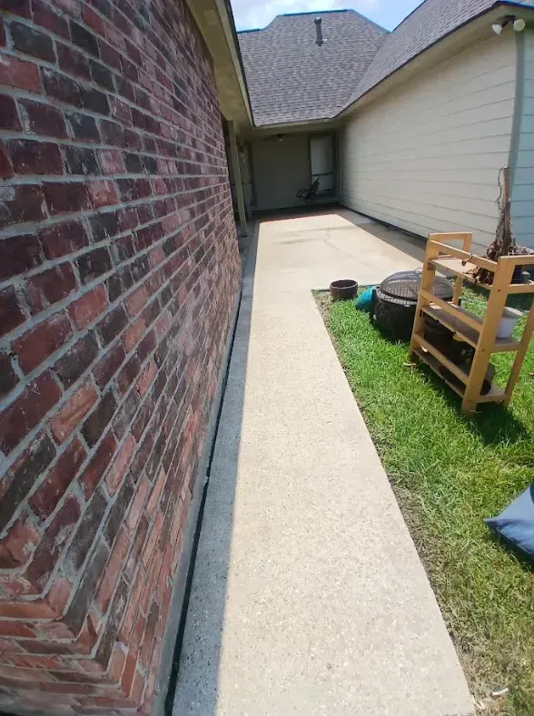 A concrete walkway leads between a brick house wall and a grassy yard with a wooden shelving unit.
