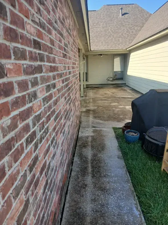A narrow concrete walkway runs between a brick wall and a patch of grass leading to a recessed patio area.