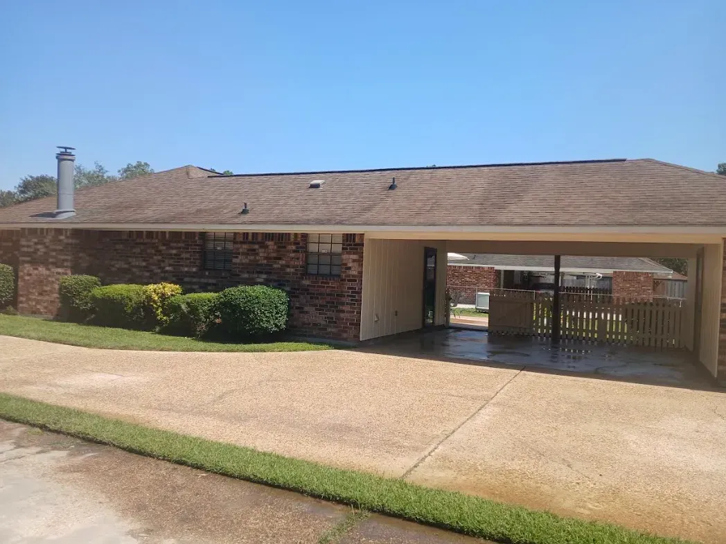 A single-story brick house with a brown roof and a large covered carport, viewed from a gravel driveway on a sunny day.