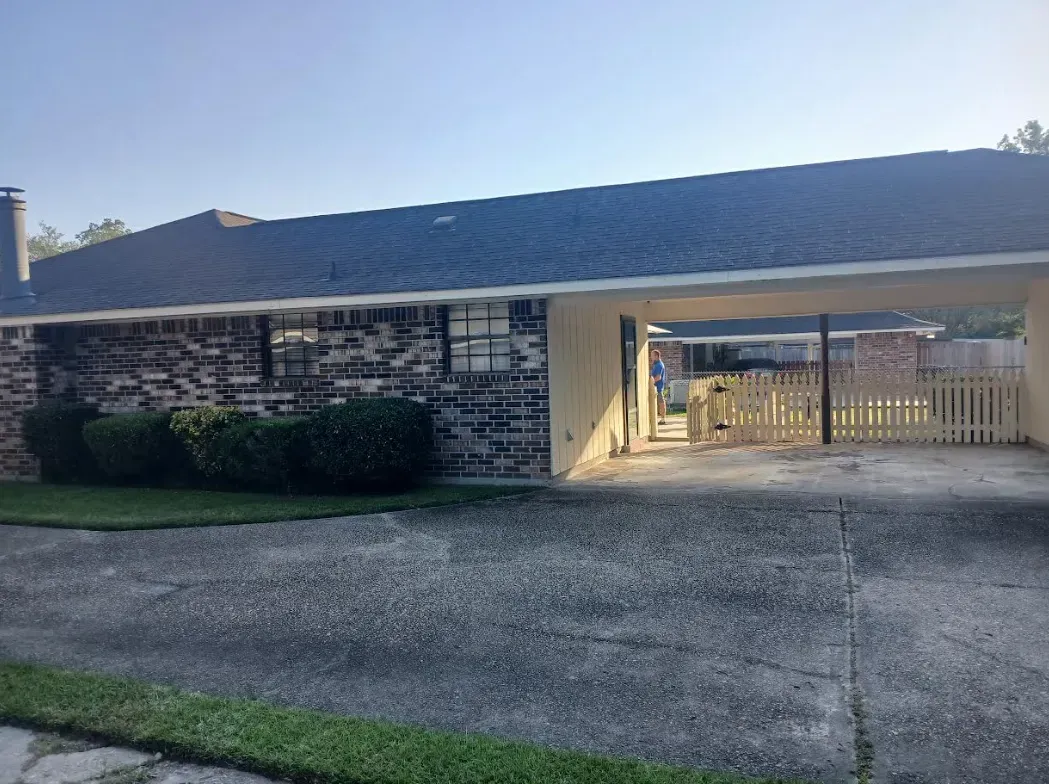 A single-story brick house with a large attached carport and a white picket fence in the driveway under a clear blue sky.