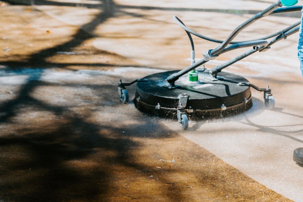 A person uses a circular surface cleaner to power wash a dirty concrete patio, revealing a clean, light-colored path.