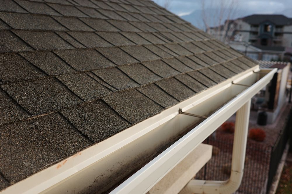 Close-up of a house roof with dark shingles and a white gutter system with an exposed screw mounting.