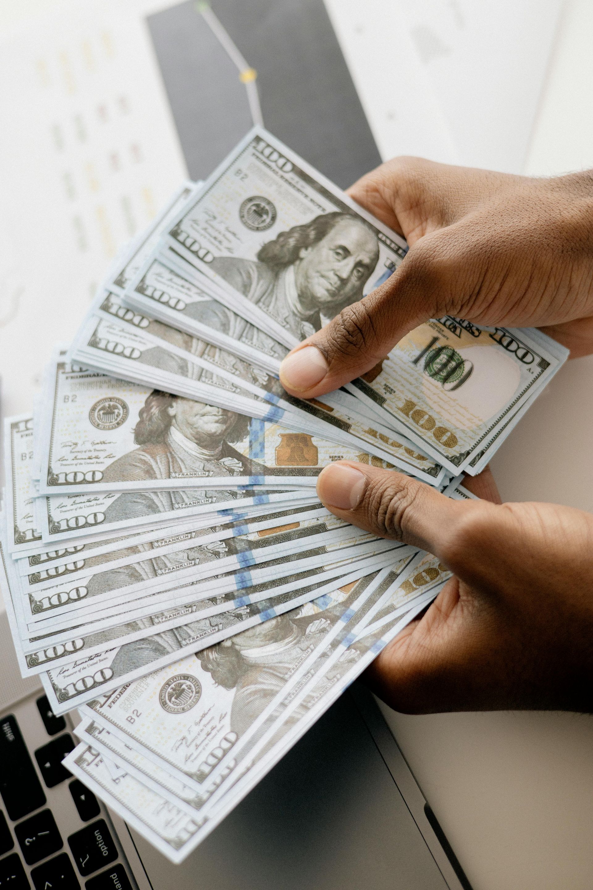 Hands holding a large stack of hundred-dollar bills near a laptop.