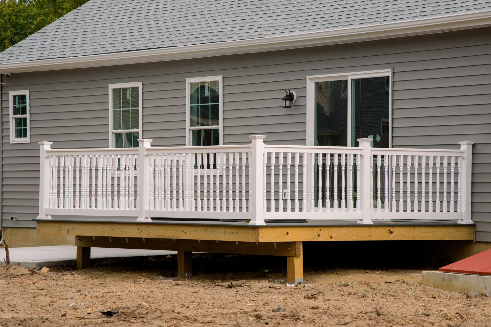 Raised wooden deck with white railing attached to a gray-sided house on a dirt lot