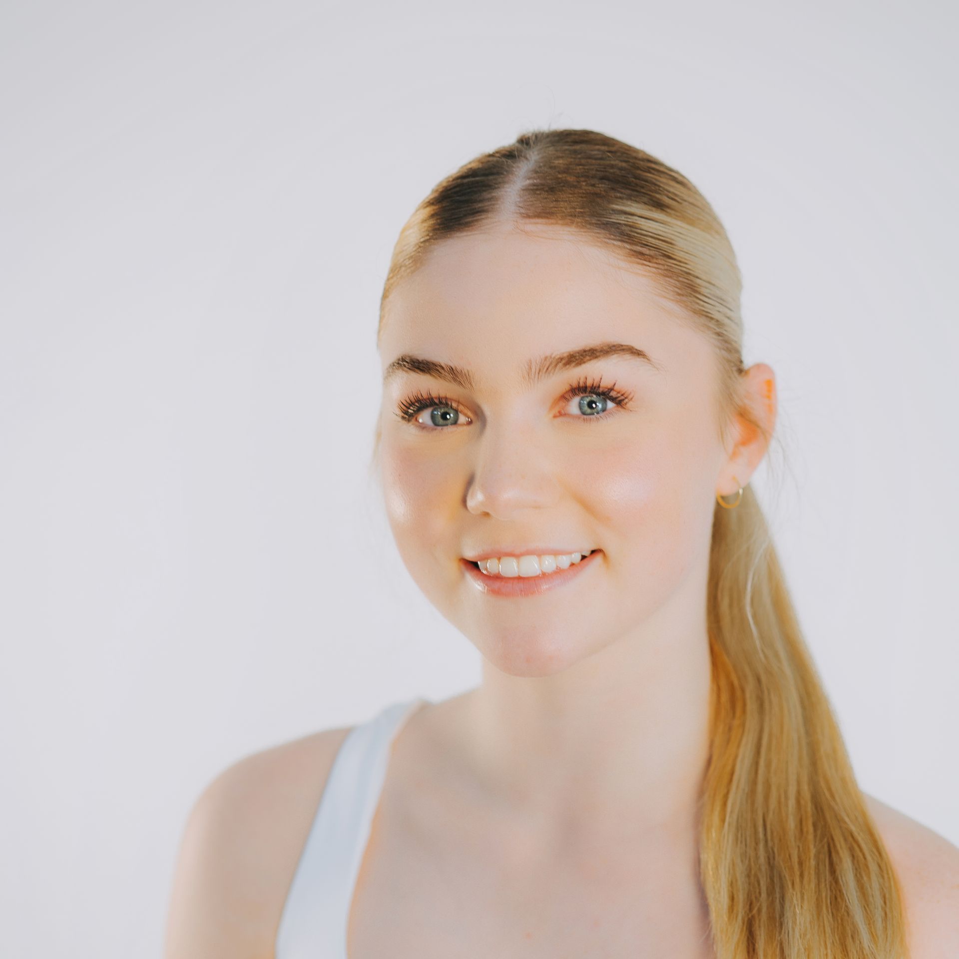A woman with long blonde hair is smiling and wearing a white tank top.