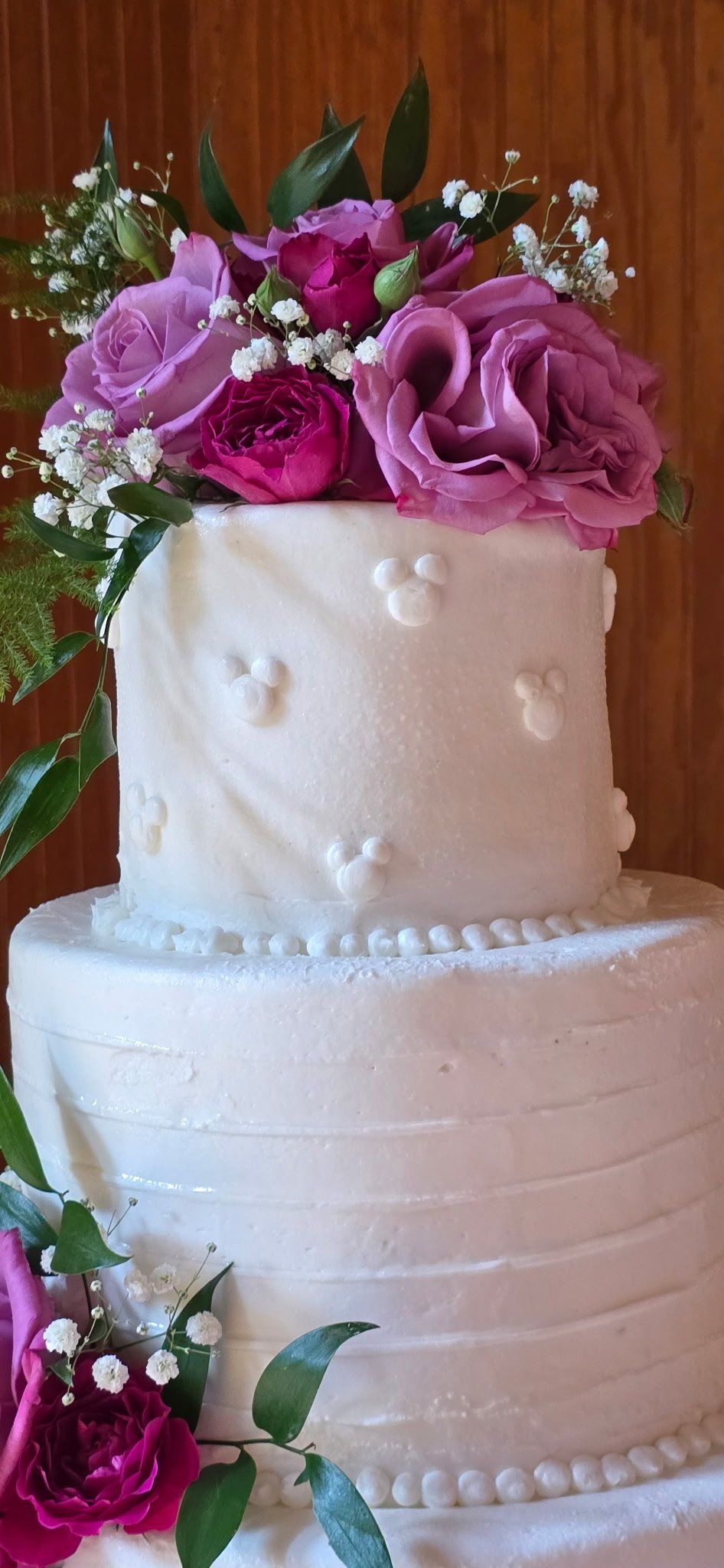 Three-tiered white cake with Mickey Mouse accents and purple flower arrangement.