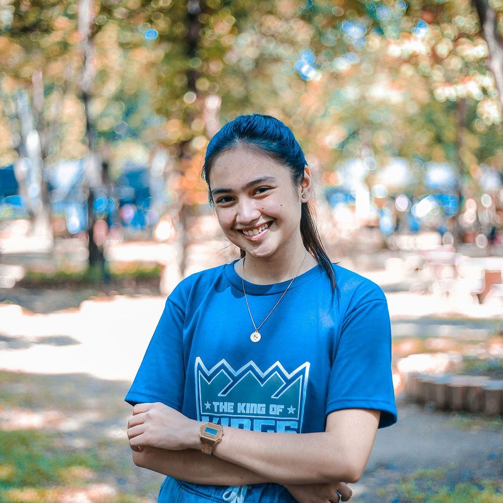 A woman wearing a blue shirt with a crown on it is standing in a park with her arms crossed and smiling.