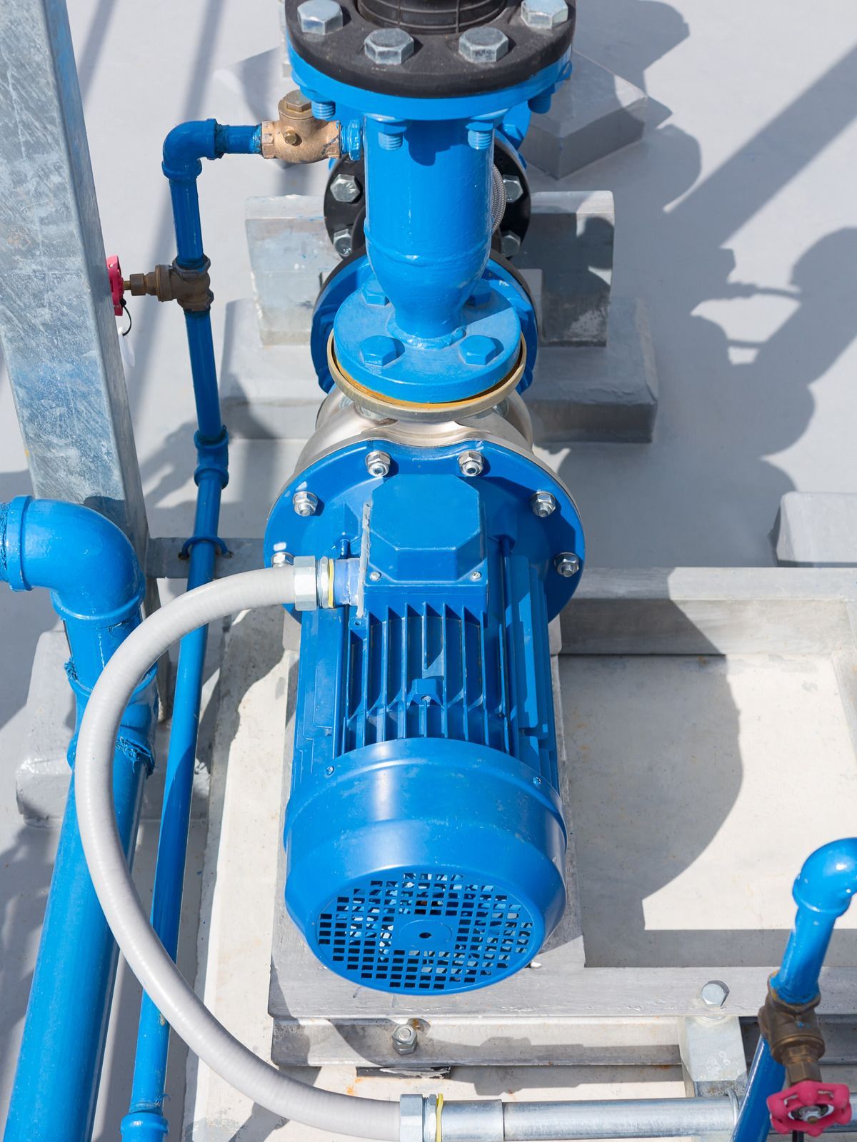 A vertical blue industrial water pump with attached plumbing, mounted on a concrete base against a light gray wall.