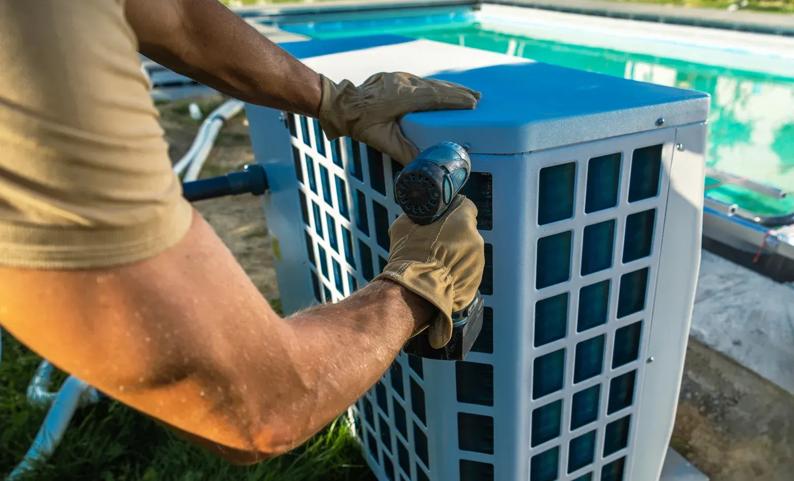 A worker wearing tan gloves uses a power drill to assemble a blue outdoor pool heat pump beside a swimming pool.