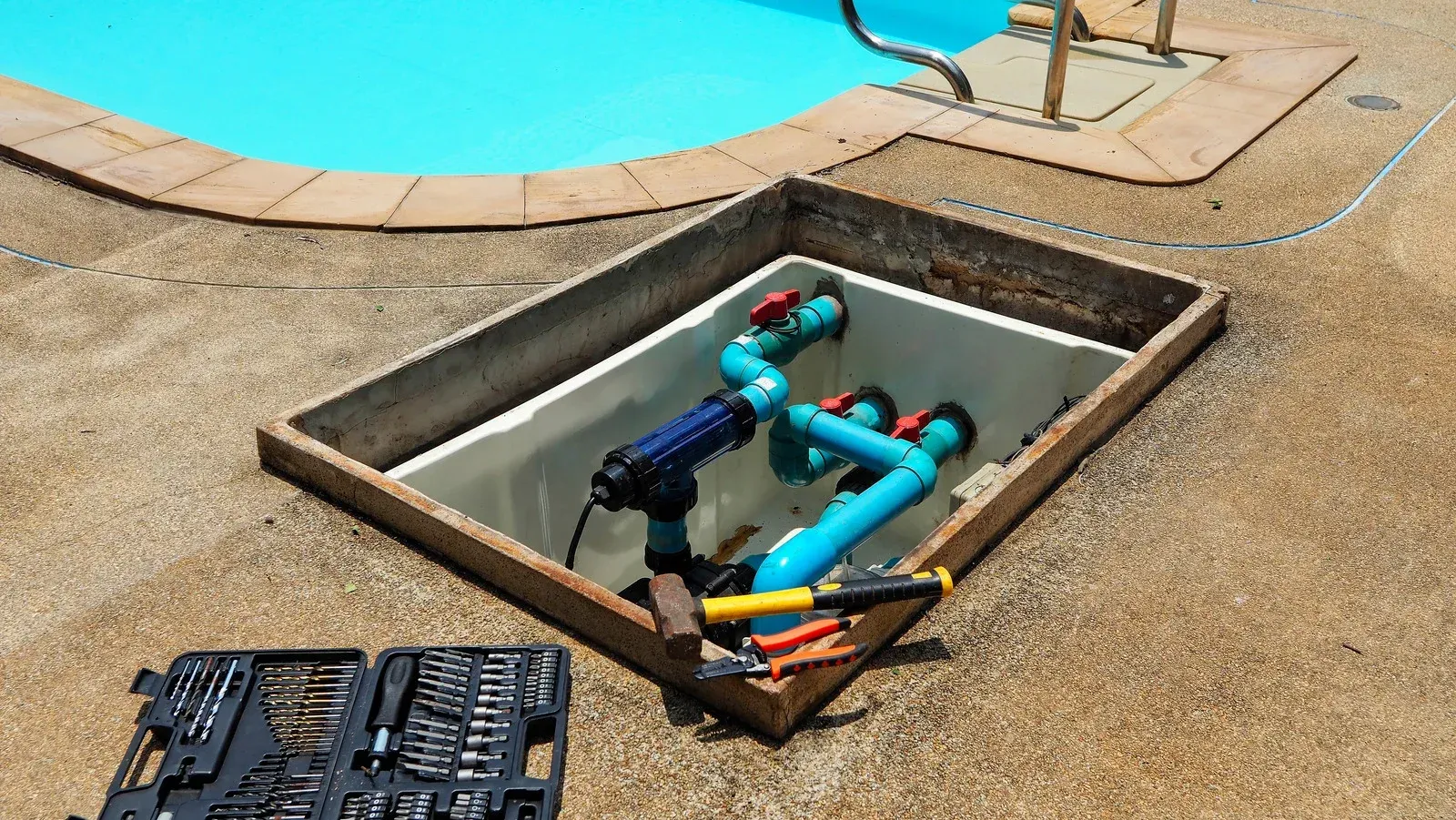 Open pool plumbing equipment pit with pipes and valves, tools in a case nearby, on a concrete pool deck.