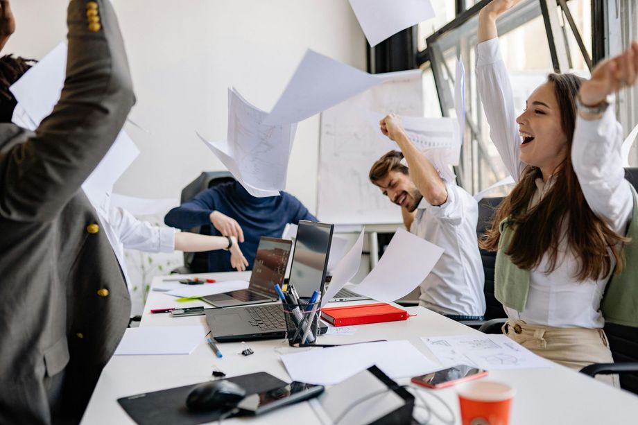 Office colleagues celebrating, throwing papers in the air. People smile, arms raised, at a desk with laptops.