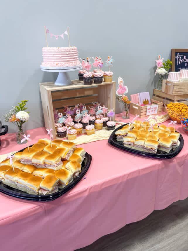 Pink-themed party food table with sliders, cupcakes, cake, and snacks.
