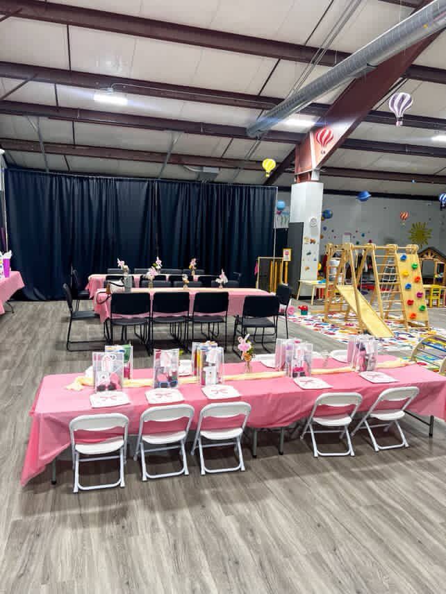 Tables set with pink tablecloths, chairs in a party setting. Background includes playground, balloons.