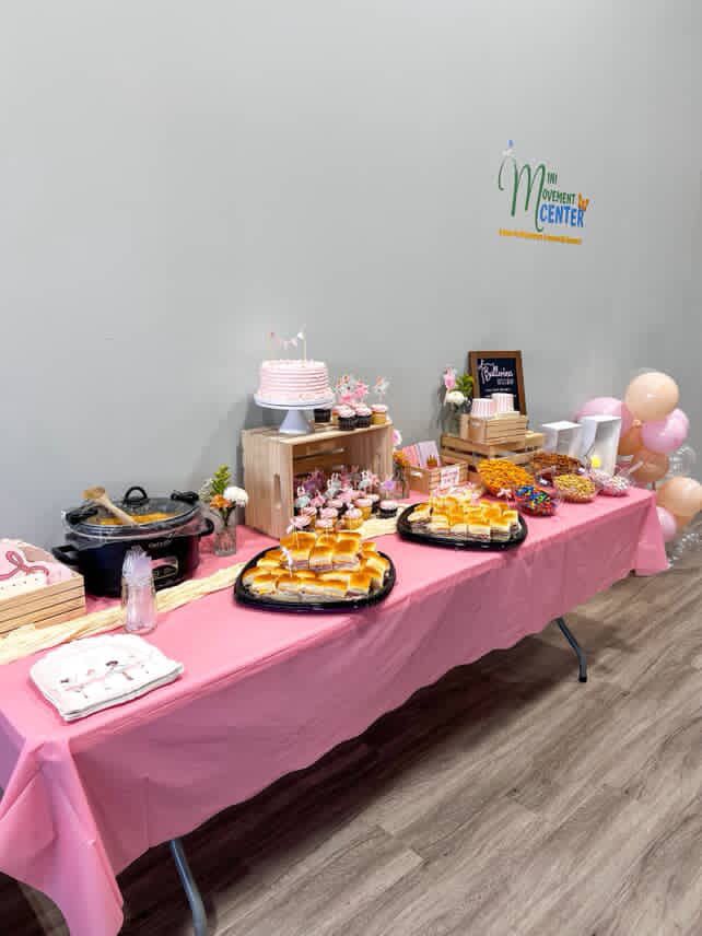 Pink-covered table with food and decorations, including cake, cupcakes, and balloons, in a room.
