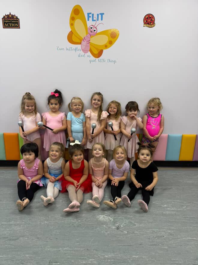 Group of children in dance attire posing in front of a butterfly mural, some holding microphones.