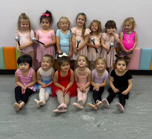 A group of young children in dance attire poses, holding props and smiling in a studio.