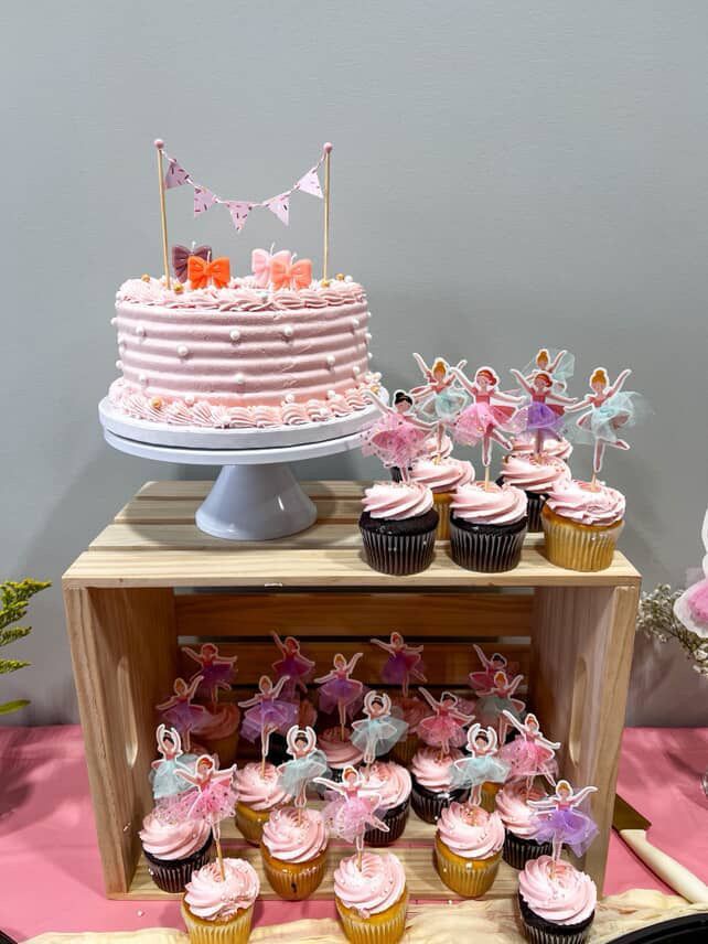 Pink ballet-themed dessert display: cake on pedestal, cupcakes in wooden crate, and toppers.