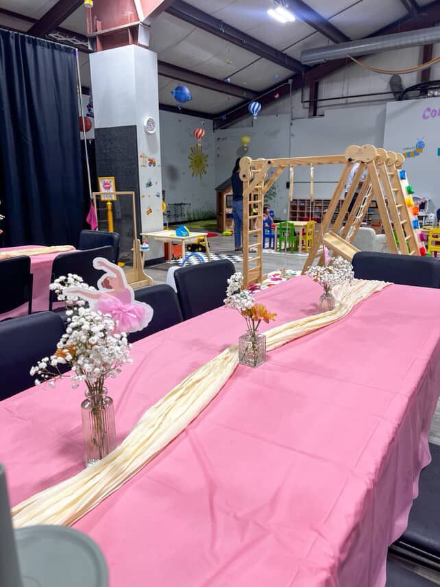 Pink table set for a party, with flowers and a wood playset in the background.