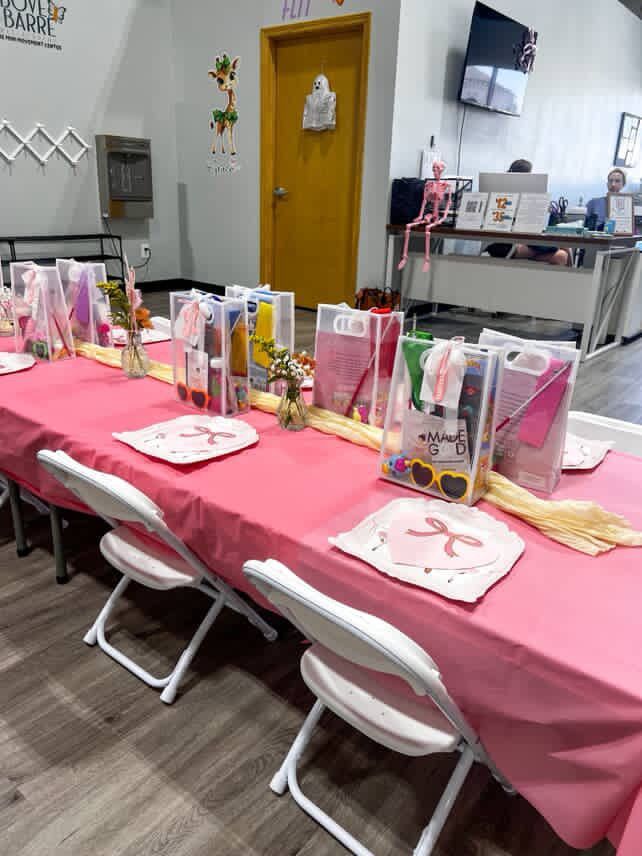 A party table set with pink tablecloth, chairs, and gift bags in a brightly lit room.